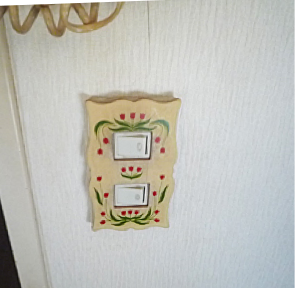 交換前の電気スイッチ。古い花柄のデザインが施された装飾プレートが設置されており、経年による汚れやくすみが全体的に見られる。