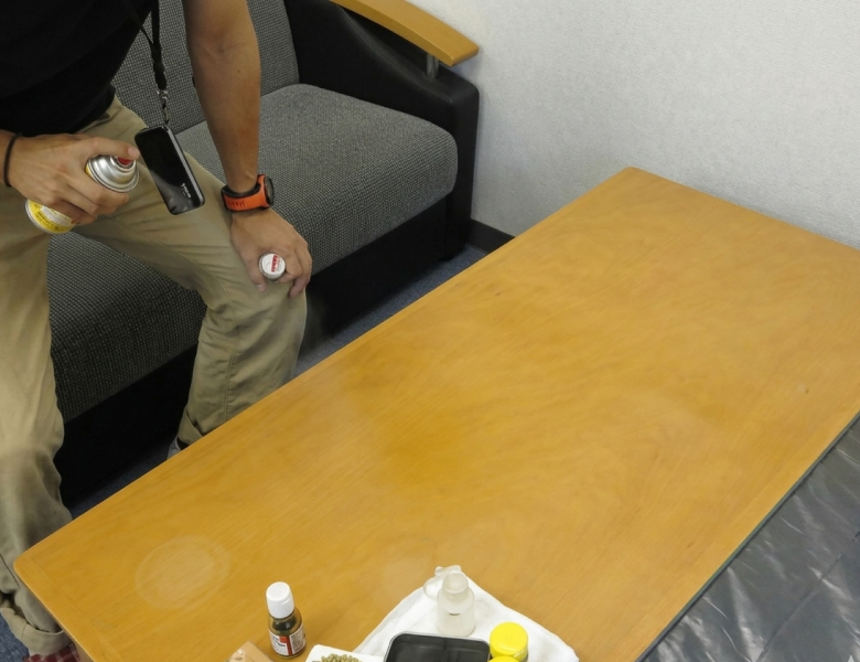 木目調のテーブルに対し、補修面を平滑にするためのスプレーを吹きかけ、周囲と馴染ませている作業風景